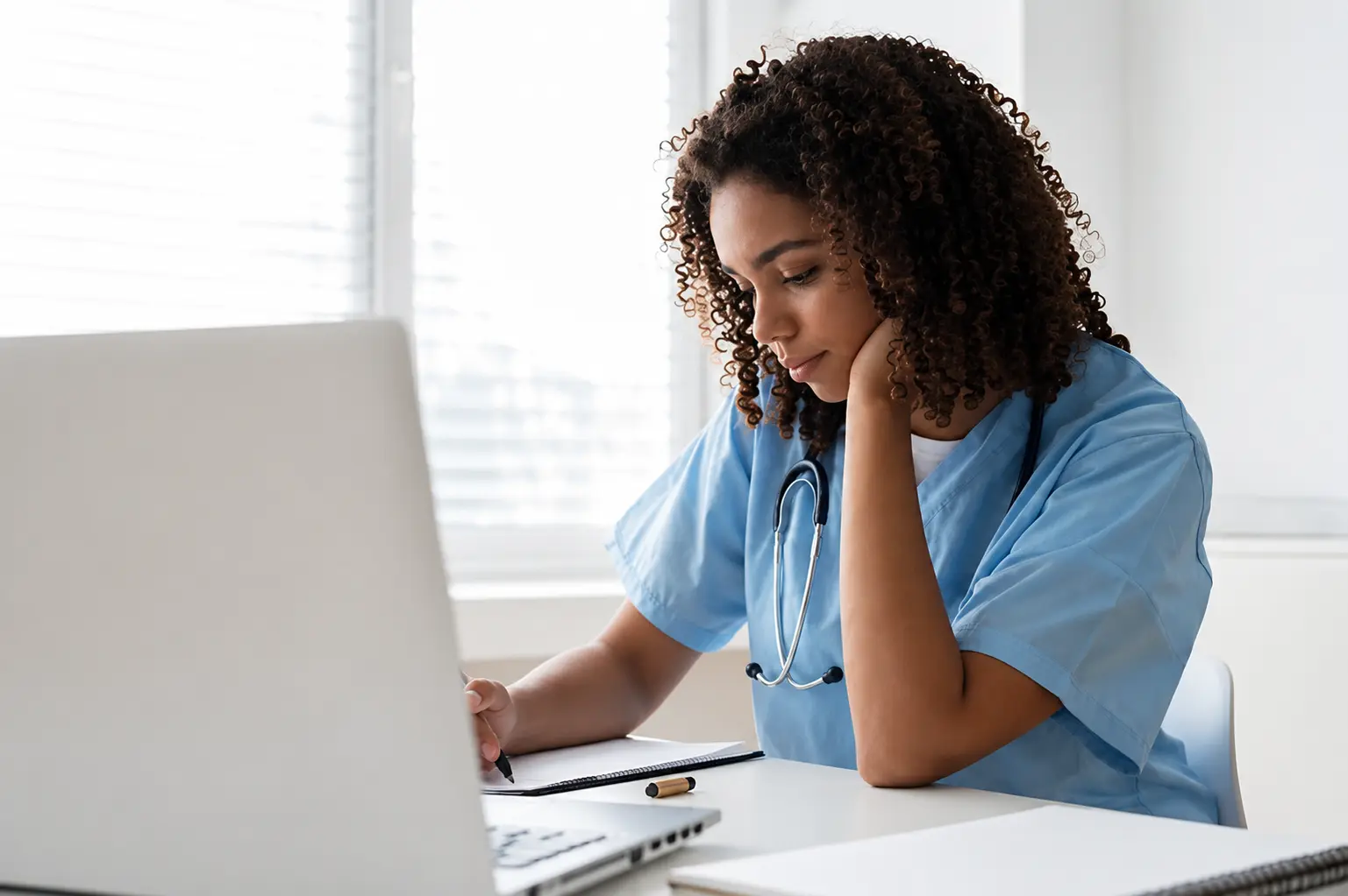 Estudante de medicina de jaleco azul e estetoscópio, estudando com caderno e notebook em uma mesa junto à janela