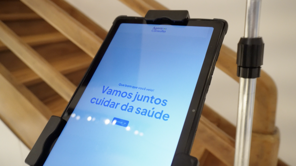 Tablet exibindo a tela inicial da plataforma Agora Consulta com a mensagem ‘Vamos juntos cuidar da saúde’, instalado em suporte em ambiente de atendimento